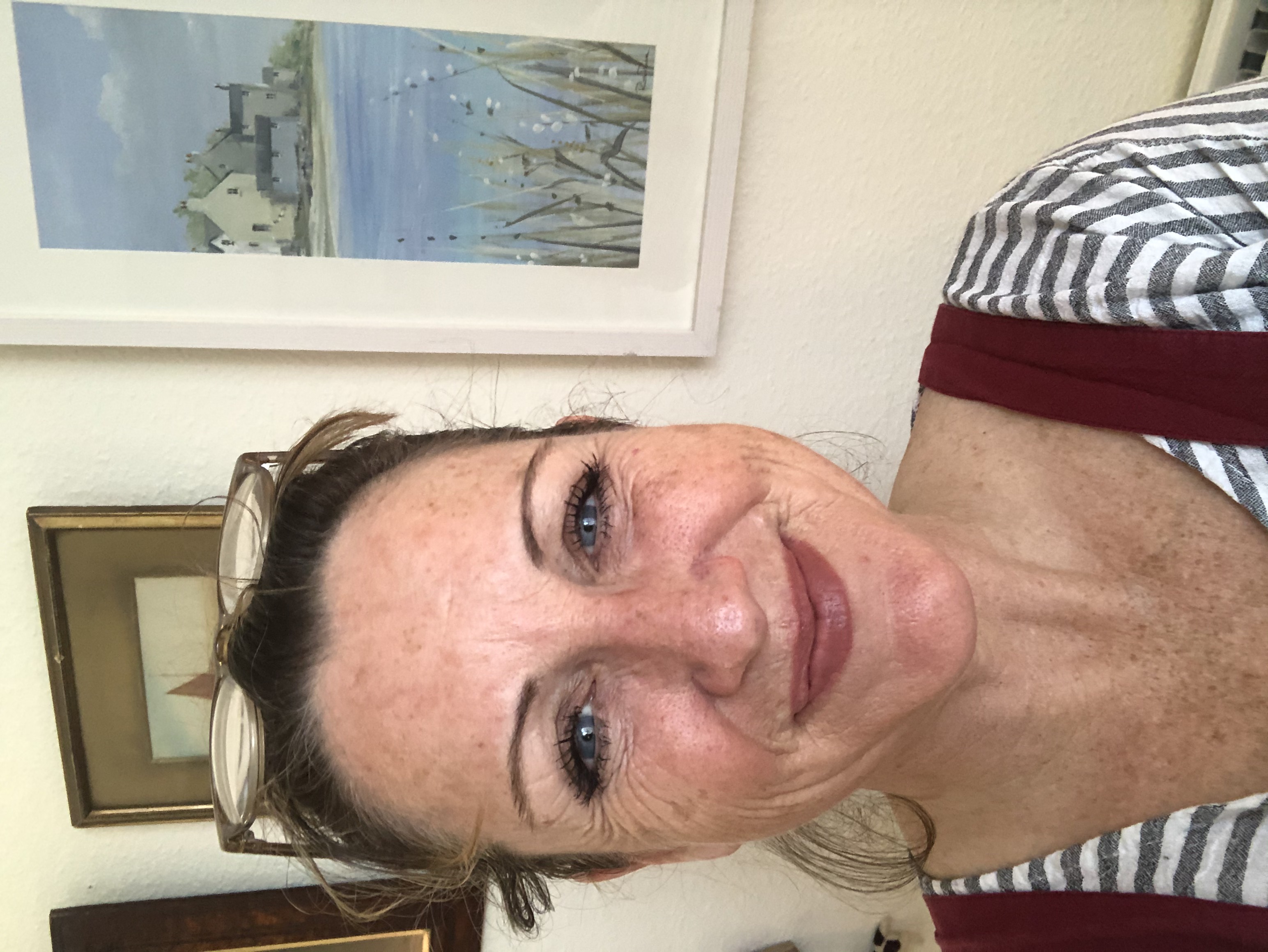 Louisa Dudley, smiling, greying dark hair up, in a red and white striped top, with framed watercolours of a harbour behind her.
