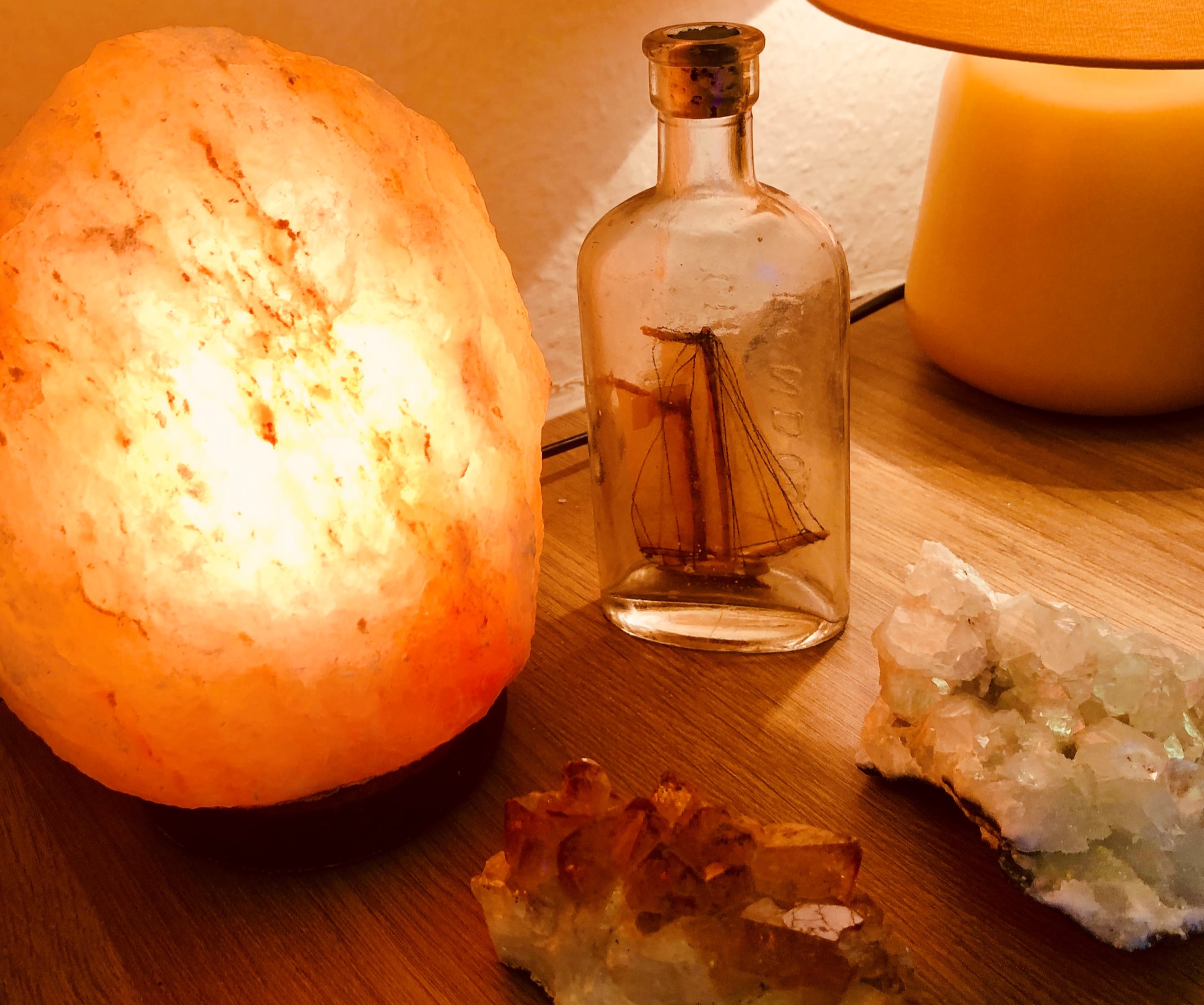 A lit Himalayan salt lamp, a small glass apothecary bottle and raw quartz and citrine clusters on a wooden table.