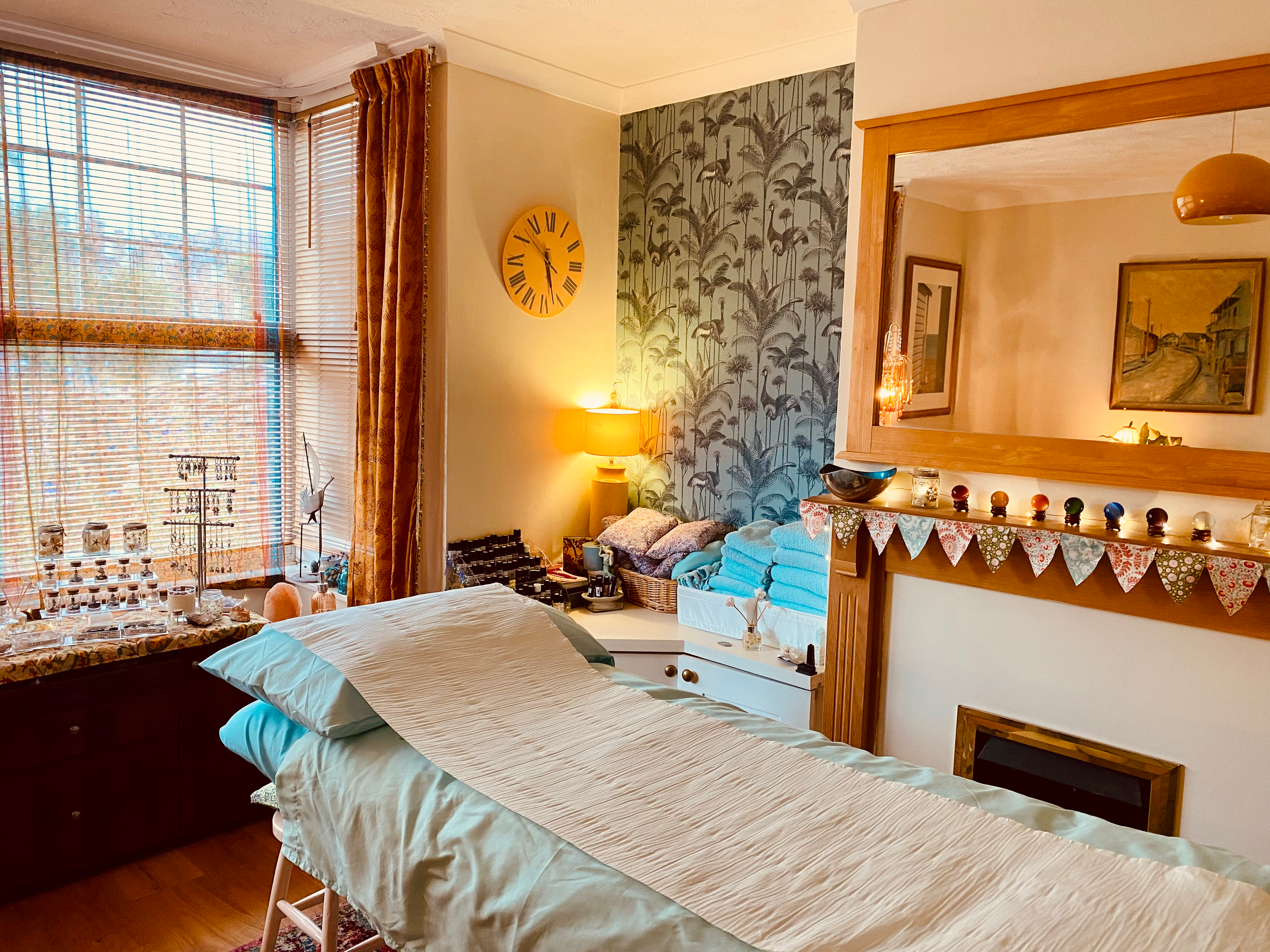Louisa's treatment room: a massage couch made up in sage-green towels under lamp light, a sash window with heavy curtains, botanical wallpaper, a mirror over the fireplace and bunting on the mantel.